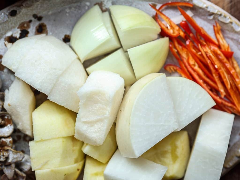 A plate with chopped ingredients including onion quarters, sliced red chili peppers, cubed white radish, and potato chunks. The items are arranged closely together, showcasing a variety of textures and colors.